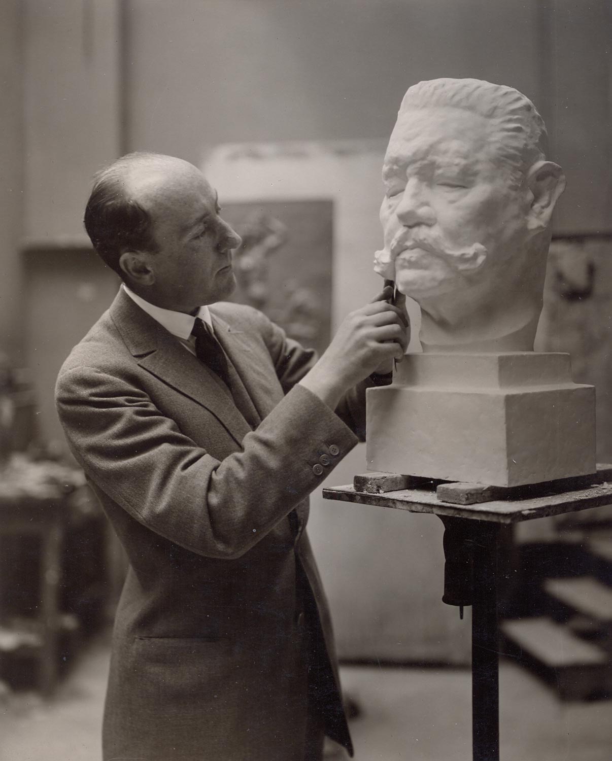 Edwin Scharff steht in seinem Atelier und arbeitet an der Hindenburgbüste. Er trägt einen Anzug und hält in der rechten Hand ein Werkzeug. Die Hindenburgbüste steht auf einem Sockel auf Augenhöhe von Scharff.