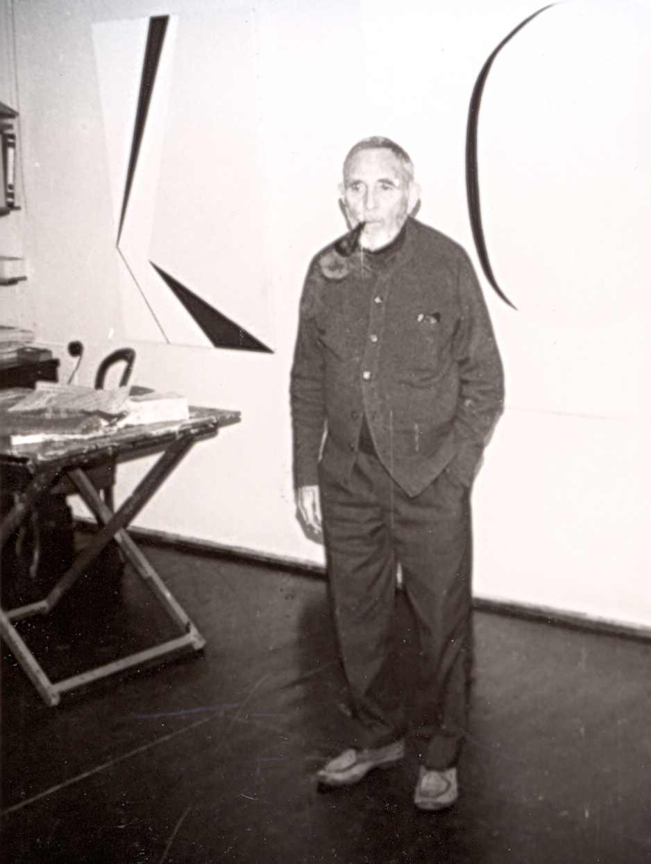Ernst Geitlinger steht in seinem Atelier. Die linke Hand hat er in die Hosentasche gesteckt. Er raucht eine Pfeife. Das Foto entstand 1971.