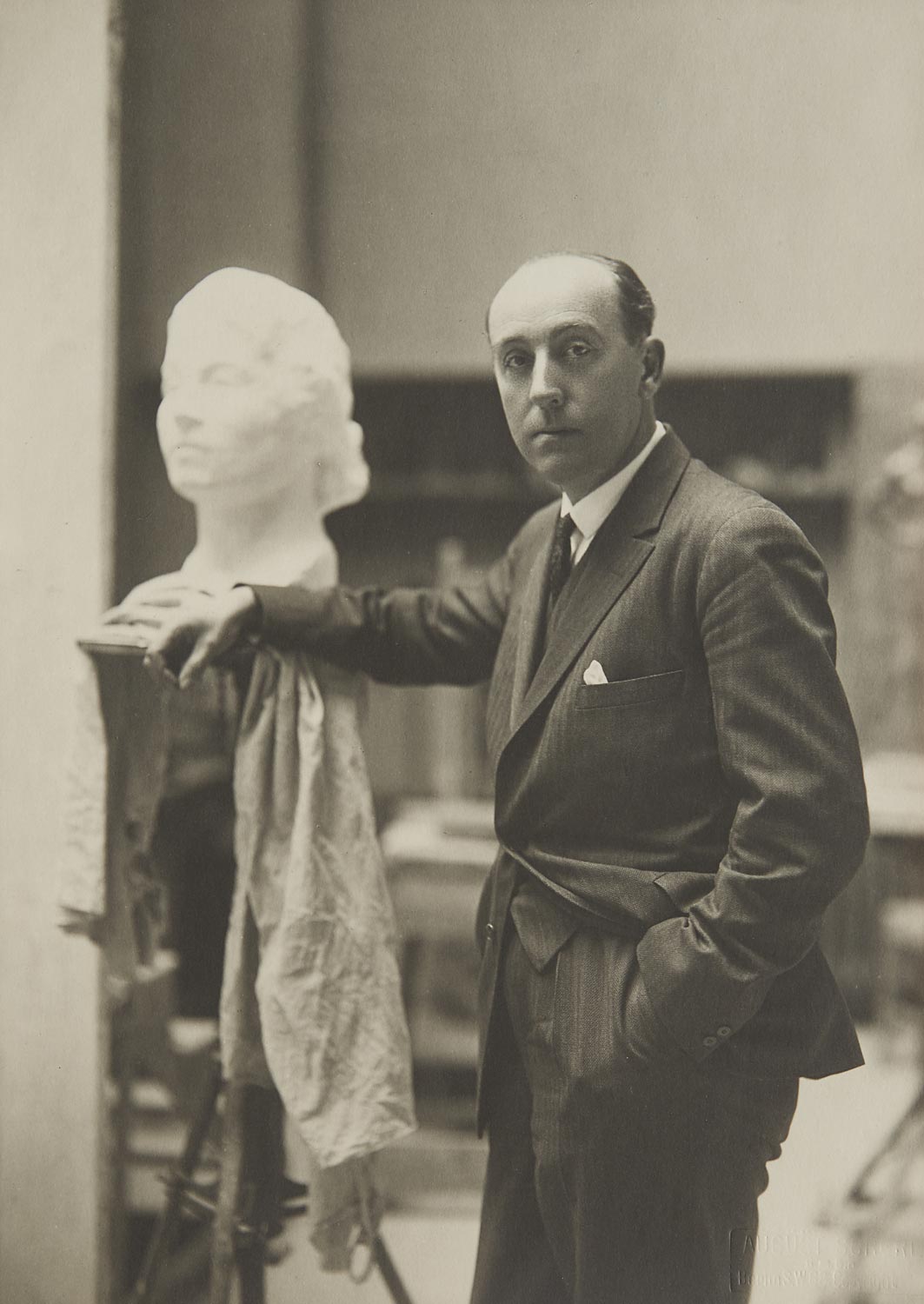 Das Foto zeigt Edwin Scharff und wurde 1929 in Berlin aufgenommen. Er blickt in die Kamera und hat seinen rechten Arm auf einen Sockel abgelegt, auf dem einen Frauenbüste steht.