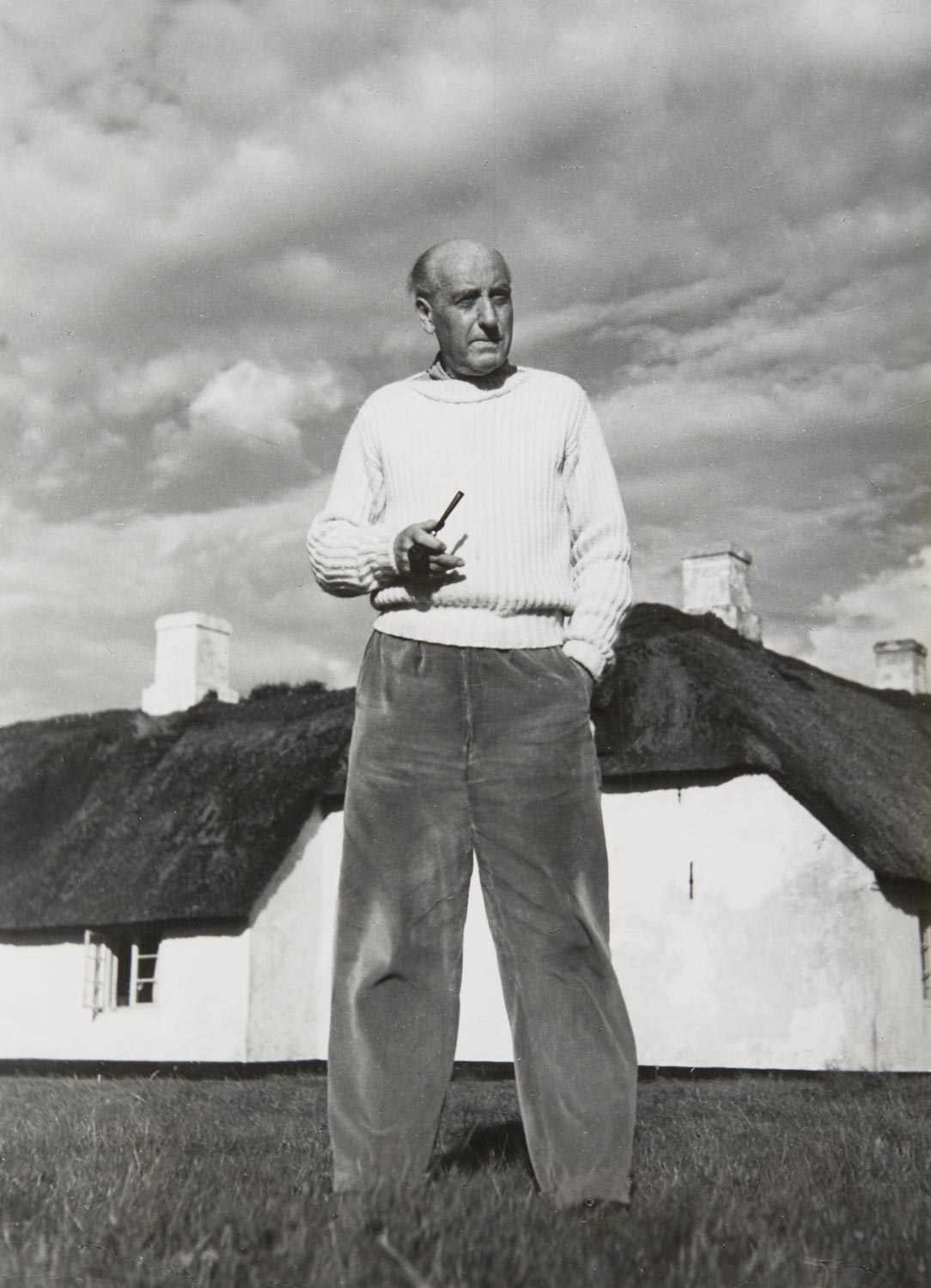 Edwin Scharff steht vor seinem Bauernhaus auf Sylt, 1950. In der rechten Hand hält er eine Pfeife, er trägt einen hellen Pullover und eine legere Hose. Im Hintergrund sieht man das Haus mit Reetdach.
