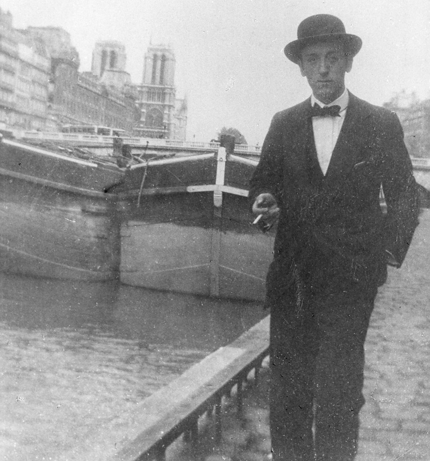 Edwin Scharff in Paris, 1912. Der Künstler blickt in die Kamera und trägt einen Anzug und Hut. In der rechten Hand hält er eine Zigarette. Er steht an einem Fluss, hinter ihm sieht man Boote, die angelegt haben.