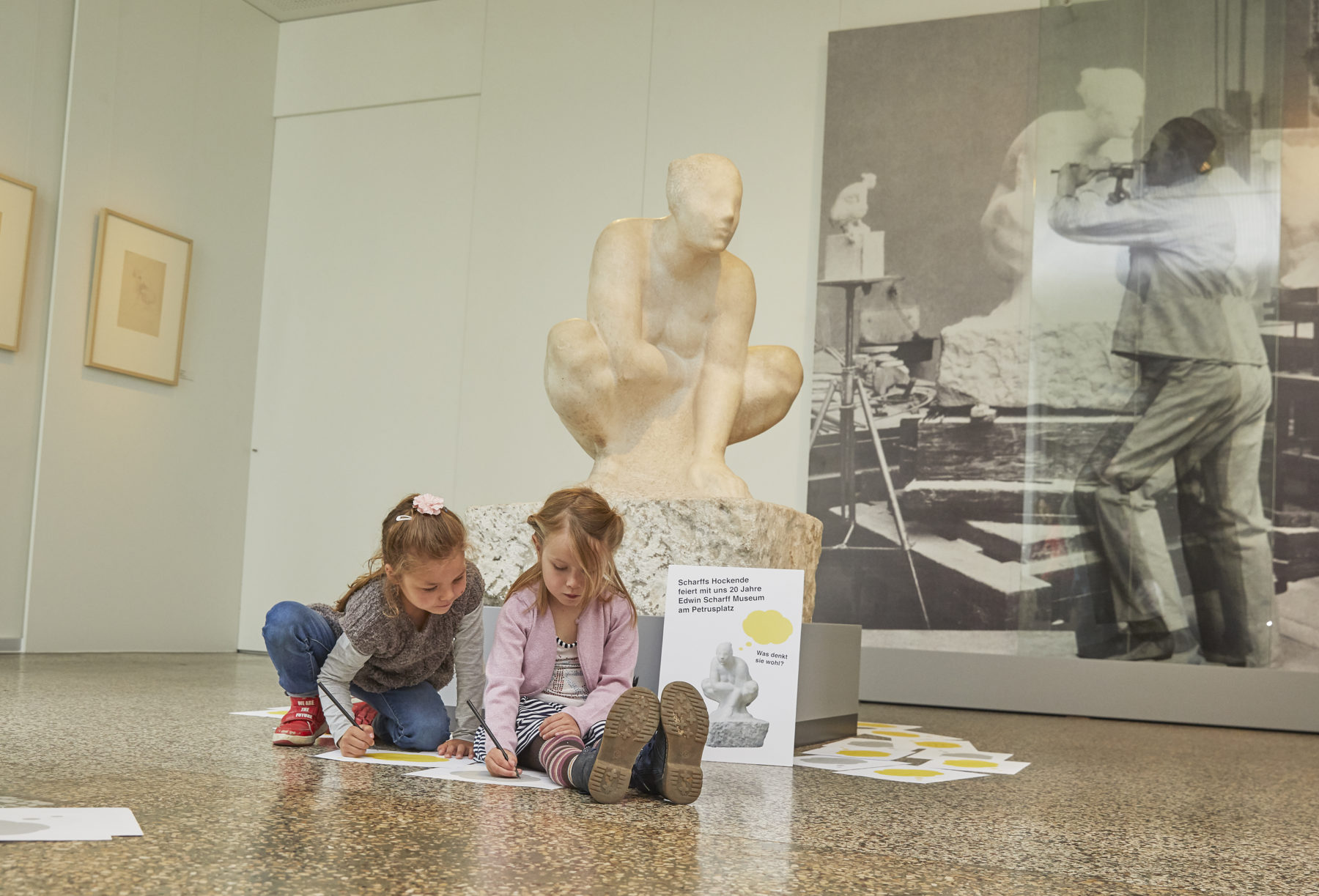Das Foto zeigt zwei Mädchen vor der Skulptur Die Hockende von Edwin Scharff. Sie sitzen auf dem Boden und nehmen an einem kreativen Angebot teil.