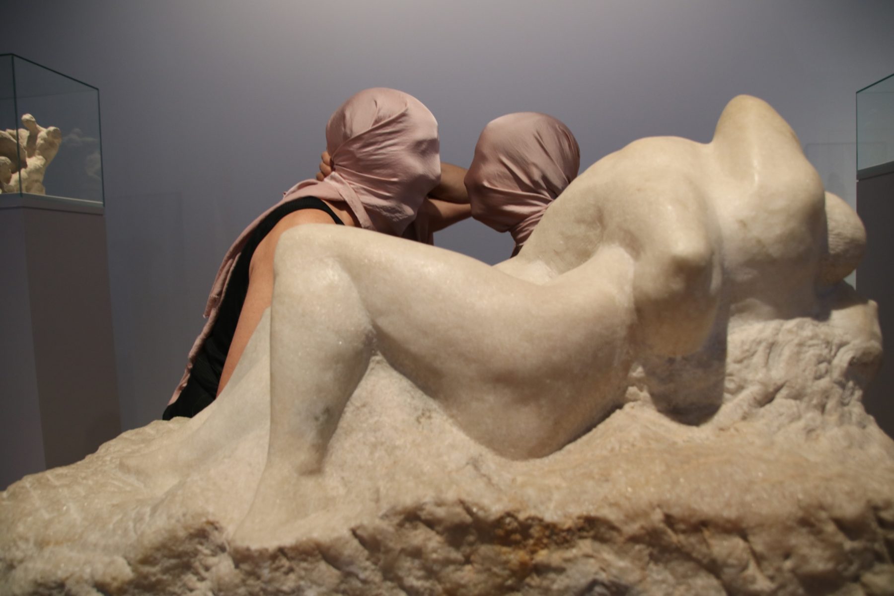 Das Foto zeigt zwei Personen, die hinter dem Kunstwerk Liebespaar von Edwin Scharff sitzen. Sie sind einander zugewandt, tragen aber zwei rosefarbene Tücher über den Kopf, sodass sie sich nicht sehen können.