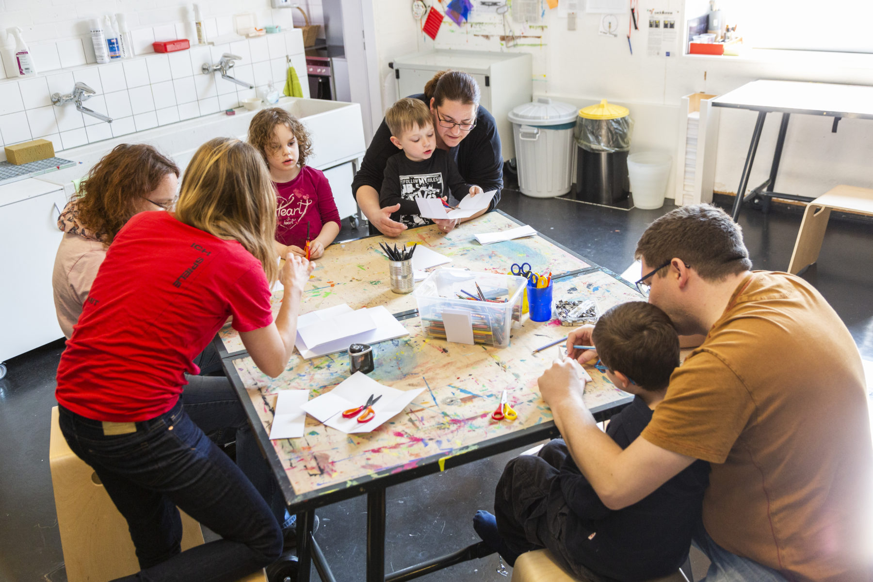 Eine Museumsmitarbeiterin erklärt Familien mit Kindern, die an einem Tisch in der Museumswerkstatt sitzen, eine kreative Aufgabe.