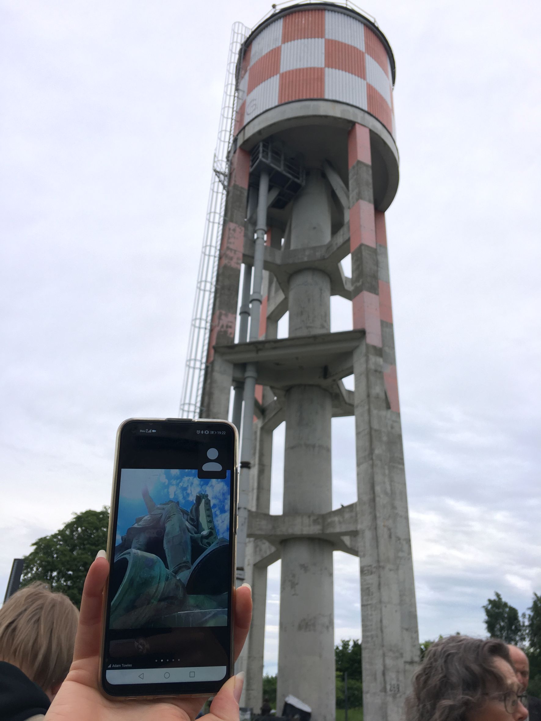 The photo shows a smartphone in the foreground with the Hermann Monument in New-Ulm, USA. The background shows Neu-Ulm water reservoir in Neu-Ulm Wiley, also a landmark of sorts.