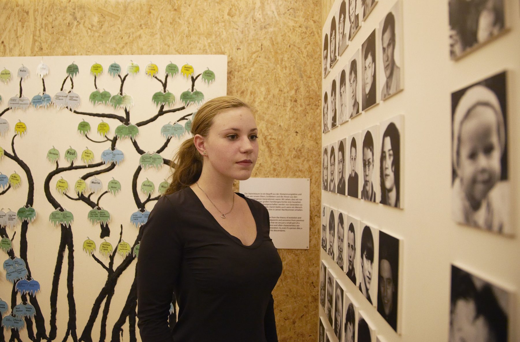 Eine junge Frau steht in einem Ausstellungsraum. Im Hintergrund an der Wand sieht man einen Baum an die Wand gemalt. Sie schaut sich Fotos verschiedener Menschen an. Es handelt sich um die Station "Galerie der Lebensalter".