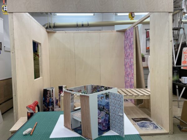 Im Vordergrund steht ein kleines, buntes Modell eines Raums. Im Hintergrund entsteht dieser Raum aus Holz, es stehen Tapeten und Arbeitsmaterial herum.