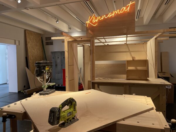 Im Vordergrund steht eine Säge auf einem Stück Holz. Im Hintergrund erkennt man einen aus Holz gebauten Raum im Raum, der in orangener Leucht-Schrift mit "Könnomat" überschrieben ist.