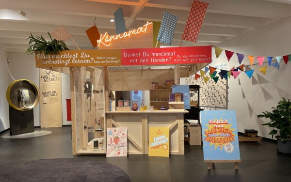 Eine Kiosk-ähnliche Holzhütte steht in einem großen Raum. Auf dem Kiosk steht in orangener Leuchtschrift "Könnomat". Drumrum ragen Fahnen aus der Hütte und auf dem Kranz ringsum stehen Fragen wie "Was möchtest du unbedingt lernen". Davor sind Plakate aufgestellt, die zum Mitmachen einladen. Im Hintergrund steht eine Telefonzelle aus den 70er Jahren.