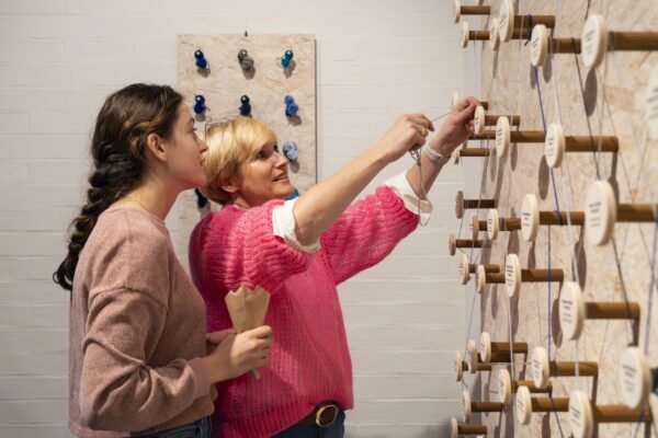 Eine Frau und eine Jugendliche stehen zusammen vor einem großen Fadendiagramm. Die Frau in pinkem Pullover legt einen Faden um ein rundes Holz-Element, auf dem eine Fähigkeit aufgedruckt ist, die sie besonders gut kann.