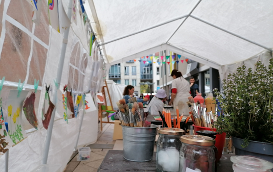Auf dem Foto ist ein Zelt zu sehen, darin sind Personen, Pinsel und Farben stehen auf dem Tisch.