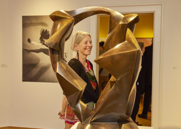 Das Bild zeigt im Vordergrund eine Skulptur in der Ausstellung "Tanz wird Kunst, Teil 2". Im Hintergrund sieht man die Kuratorin Ina Ewers-Schultz lächelnd.