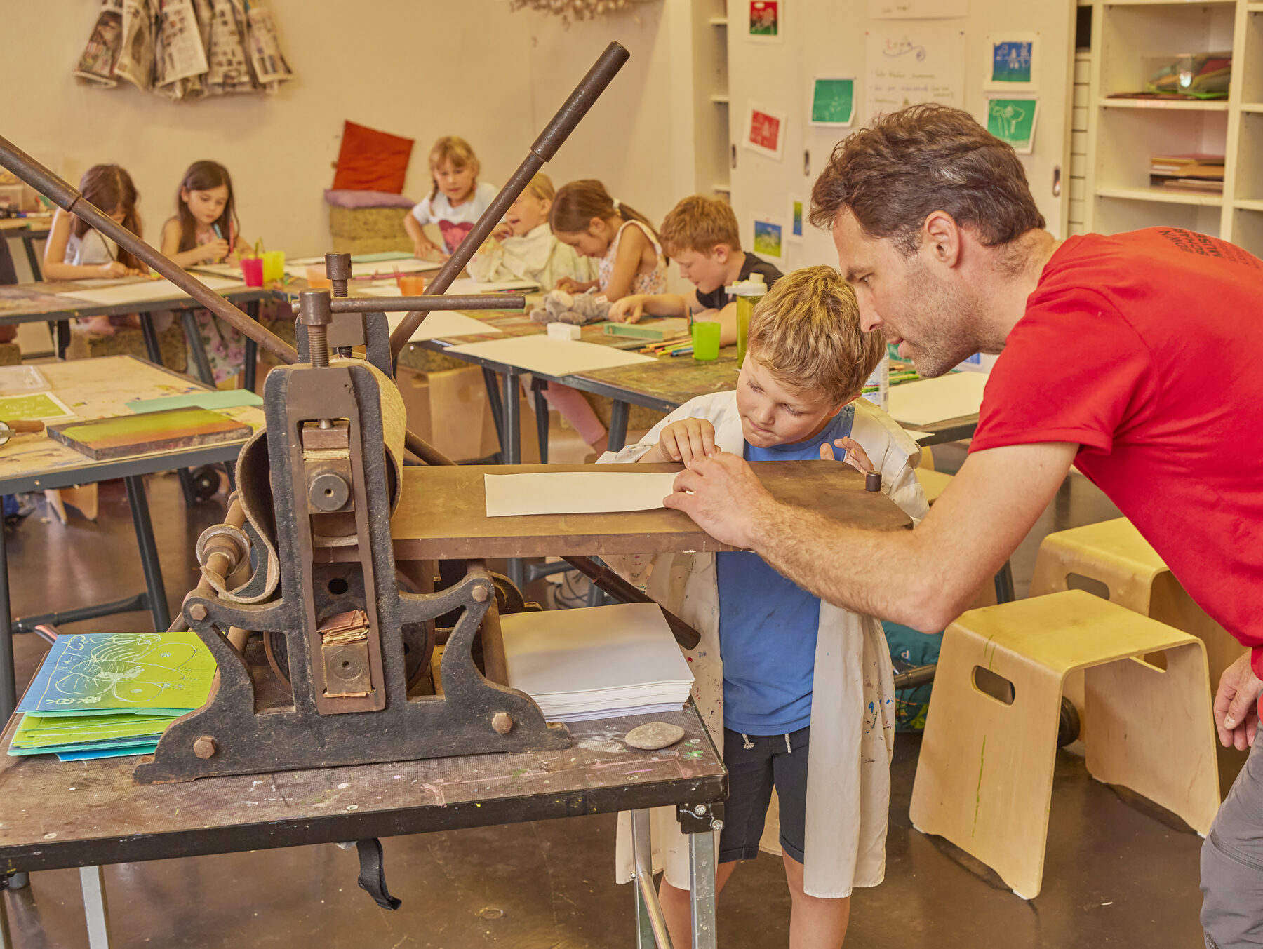 Das Bild zeigt einen Blick in die Museumswerkstatt. Kinder sitzen an Tischen und malen, im Vordergrund hilft ein Museumspädagoge einem Kind beim Druck eines Bildes.