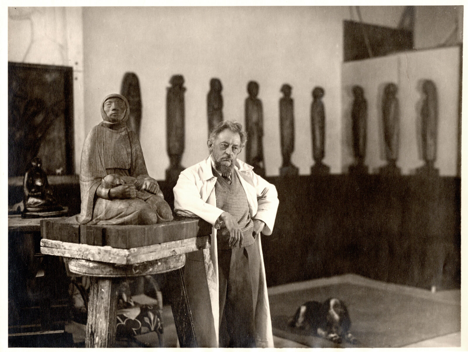 Die Schwarzweiß-Fotografie zeigt den Künstler Ernst Barlach in einem Atelier. Er lehnt in einem weißen Kittel neben seiner Holzskulptur mit dem Titel "Mutter und Kind". 